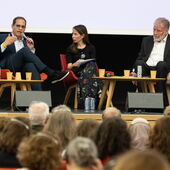 Drei Personen sitzen auf dem Podium und diskutieren, im Vordergrund sind unscharf die Hinterköpfe des Auditoriums zu sehen.