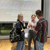 Drei Personen stehen vor der Leinwand und unterhalten sich, einer hält ein Glas Sekt in der Hand.