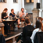 The speakers sit on a podium in front of an audience in a courtyard. Sébastien François speaks into a microphone.
