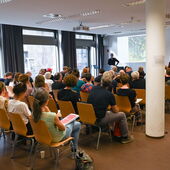 A full seminar room, the audience looks at the speaker at the lectern. Large windows face the street on the left.