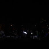 Auf einer Straßenkreuzung im Dunkel sind schemenhaft ein schwach beleuchteter Tisch, Hocker und auf ihnen sitzende Menschen zu erkennen.