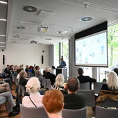 From the corner of the room, well-filled rows of audience members can be seen arranged in a semicircle, Martin Germer stands at the front of the podium and speaks.