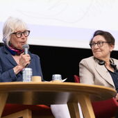 Zwei Personen sitzen auf einem Podium, die Person links spricht ins Mikro.