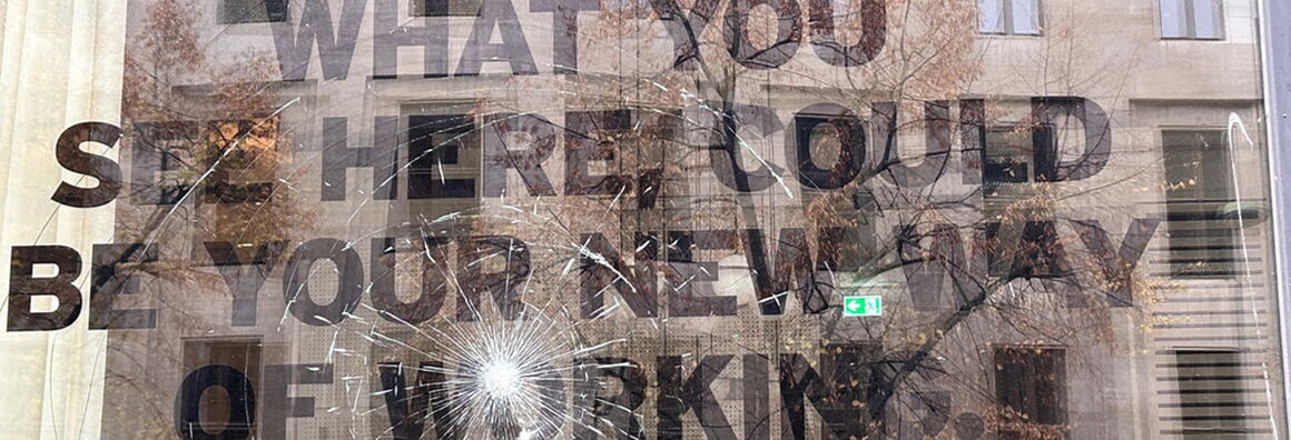 Auf einem eingeschlagenen Fenster steht "WHAT YOU SEE HERE COULD BY YOUR NEW WAY OF WORKING". Im Fenster spiegelt sich das gegenüberliegende Haus.