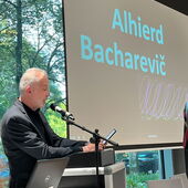 Alhierd Bacharevič stands at the lectern and reads from his e-book reader, his name displayed on the screen next to him.