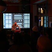 Der Musiker Adi Gelbart tätigt Einstellungen an einem Mischpult. Hinter ihm ist eine Leinwand zu sehen, welche eine Fotografie von Kabelsteckern zeigt.