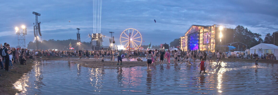 Szene vom Festival Przystanek Woodstock. Im Abendlicht ist eine große Pfütze zu sehen, in der Menschen tanzen und spielen. Hinten rechts ist eine hell beleuchtete Bühne, in der Mitte ein Riesenrad zu sehen.