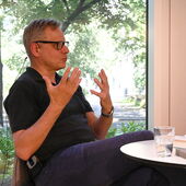 Georg M. Oswald  sitzt an einem weißen Bistrotisch und spricht gestikulierend.