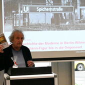 Gert Eckel stands at the lectern and holds up a copy of his novel "Unter dem Regenbogen".