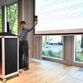 Susanne Bauer stands behind the lectern and holds a flyer towards the audience.
