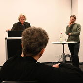 Eine Person mit schulterlangem grauen Haar, der Moderator Ernst Müller, steht am Rednerpult. Links von ihm steht der Preisträger Georg Simmerl an einem Stehtisch.