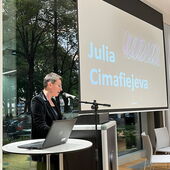 Julia Cimafiejeva steht am Rednerpult und spricht ins Mikrofon. Neben ihr auf der Leinwand steht ihr Name.