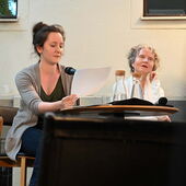 Anja Schenk und Martina Stemberger sitzen auf einem Podium, Anja Schenk hält ein Paper in der Hand.
