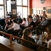 In einem hellen Raum sitzen Personen auf Stühlen und unterhalten sich, im Hintergrund links ist eine DJ zu sehen.