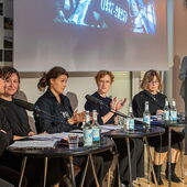 Five people are sitting on a podium and applauding. To their right, one person is standing at a lectern and speaking into a microphone.