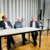 Michael Hofmann, Johannes Thorbecke and Arnd Beise sit at a table and talk. In the background are music stands in front of a light gray curtain.