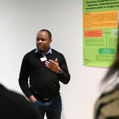 Giresse Teikeu steht vor einem Poster zum Thema »Deutsches Kaiserreich und koloniale Migration« und spricht, in Richtung Publikum gestikulierend.