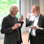 Ernst Müller and Gerwin Zohlen stand in front of a window behind which plants can be seen. Müller is talking, Zohlen is drinking from a white cup.