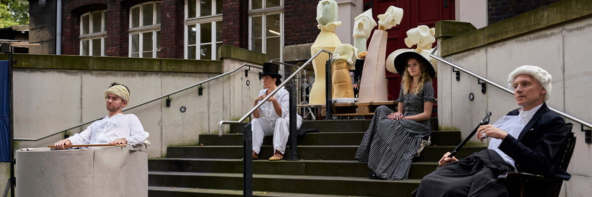 Four actors in historicising costumes sit on a staircase in front of a brick building, behind them stand various figures made of yellow and pink foam.