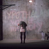 Eine Frau mit langem weißem Haar und glitzerndem Oberteil beugt sich vor einer beschriebenen Betonwand vor. An der Wand steht »Hypothalamus wall of memory«. Neben ihr steht ein kleines gebasteltes Einhorn.