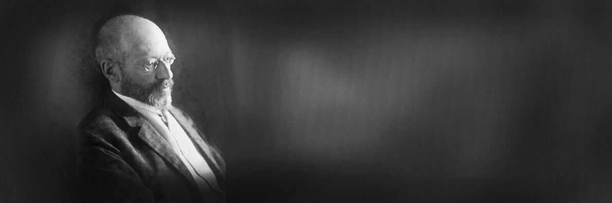 Schwarz-weiß-Bild von Georg Simmel vor schwarz-grauem Hintergrund. Simmel ist vom Kopf bis zur Mitte des Oberkörpers am linken Bildrand zu sehen und schaut zum rechten unteren Bildrand.