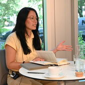 Khuê Pham sitzt mit einem aufgeschlagenen Buch an einem weißen Bistrotisch mit Tasse, Gläsern und Karaffe und spricht.