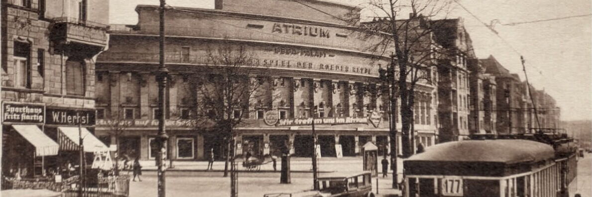 Schwarz-weiß-Foto: Historische Straßenansicht mit einem geschwungenen Eckgebäude mit der Aufschrift "Atrium" im Zentrum, darum herum Läden, eine Straßenbahn und Passant*innen.