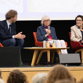Drei Personen sitzen auf einem Podium, die Person in der Mitte spricht ins Mikro.