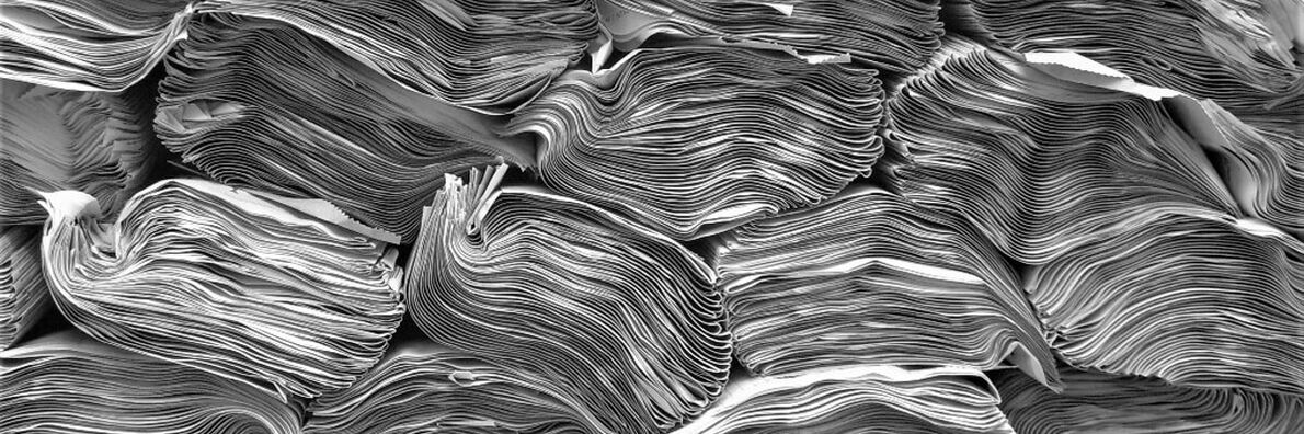 Schwarz-weißes Foto von mehreren sehr dicken, eng zusammenliegenden Zeitschriftenstapeln aus der Seitenansicht. Zusammen ergeben sie ein gewelltes Muster.