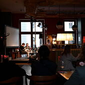 Im Hintergrund diskutieren Elias Hirschl und Hanna Hamel auf einem Podium. Im Vordergrund ist Publikum erkennbar.