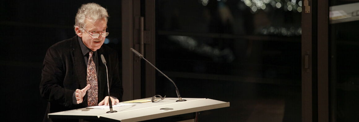 Hans-Ulrich Gumbrecht stands at the lectern, brightly lit in a dark room, and speaks.