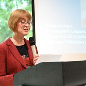 Irmela Marei Krüger-Fürhoff steht am Rednerpult und spricht ins Mikrofon. Rechts von ihr ist ein Teil ihrer Präsentation zu sehen.