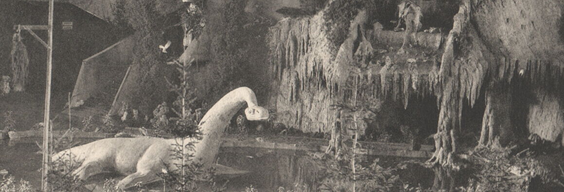 Historic black-and-white staged scene of a prehistoric landscape with a dinosaur model among artificial rocks, fir trees, and a small pond inside an indoor setting.