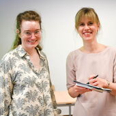 Hanna Hamel and Pola Groß stand next to each other and smile warmly at the camera. Pola Groß holds a book, paper and a pen in her hands.