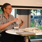 Kenah Cusanit sitzt an einem weißen Bistrotisch und gestikuliert, vor ihr liegt ein aufgeschlagenes Buch.