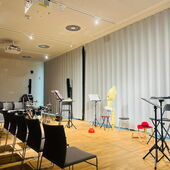 In front of a gray curtain are several stools, music stands and theater props. Opposite to the left are empty rows of chairs.