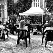 In einem kleinen Garten sitzen Menschen auf Gartenstühlen. Sie sind von hinten zu sehen und schauen auf ein kleines Zelt mit einer Bühne.