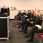 Eine Person im schwarzen Kleid spricht am Rednerpult. Vor ihr sitzen mehrere Personen, einer davon ist der Preisträger Georg Simmerl.