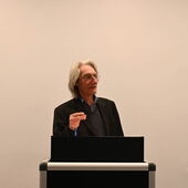 Eine Person mit schulterlangem grauen Haar, der Moderator Ernst Müller, steht am Rednerpult.