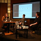 Authors Philipp Schönthaler and Hannes Bajohr and moderator Morten Paul sit on a podium. Schönthaler is bent over a laptop, Bajohr and Schönthaler turn away and look at a large screen behind them.