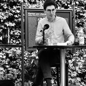 Juan S. Guse sitzt auf einer kleinen Bühne und spricht ins Publikum.