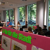 Ein voller Veranstaltungsraum. Im Vordergrund auf einer Tafel mit Zeitungsausschnitten steht »Li-Be für die Stadt«, im Hintergrund sitzen Khuê Pham und Janika Gelinek.