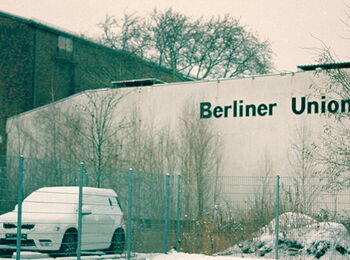 Buildings of the Berlin Union Film in winter, with a snow-covered car and bare trees in the foreground.