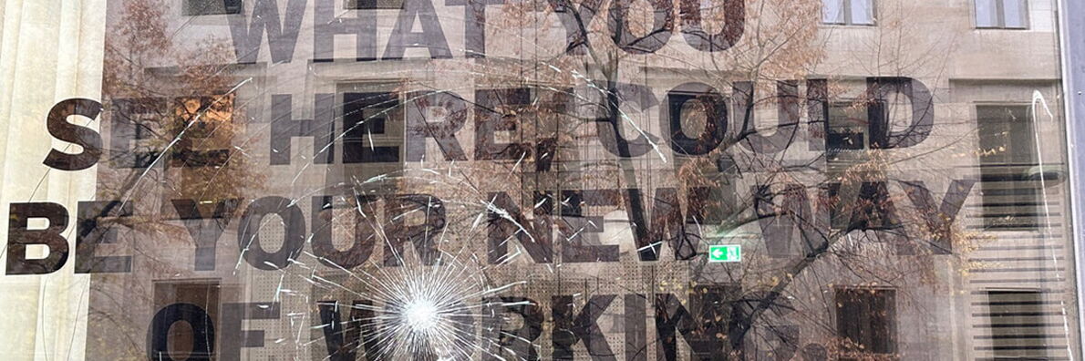 “WHAT YOU SEE HERE COULD BY YOUR NEW WAY OF WORKING” is written on a smashed window. The house opposite is reflected in the window.