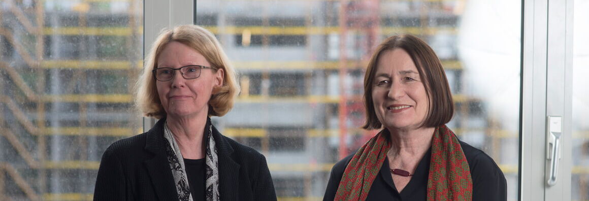 Franziska Thun-Hohenstein and Irina Scherbakowa stand side by side in front of a window, behind which a construction site can be seen, and smile at the camera.