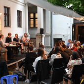 Die Sprecher*innen sitzen auf einem Podium vor Publikum in einem Innenhof. Sébastien François spricht in ein Mikrofon.