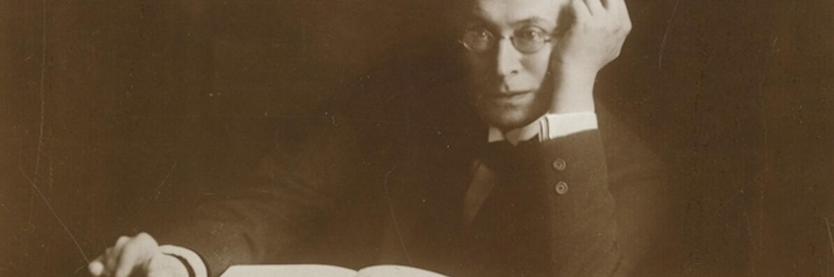 Photo of Karl Kraus. He sits at the table in a suit and glasses and looks into the camera. On the table lies an open book and a closed notebook next to it. Kraus rests one elbow on the table, his hand on his temple.
