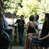 Several people stand in the Volkspark Hasenheide and listen to Cord Riechelmann's guided tour using headsets. The focus is on the author Tabea Hertzog, who has her bicycle with her.