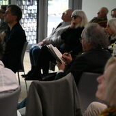 A gentleman with chin-length gray hair can be seen in the audience from diagonally behind, making handwritten notes in a notebook.