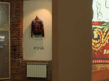 An exhibition space featuring photographs, a piece of outerwear, and a film still of a person in a colorful dress on a bed.